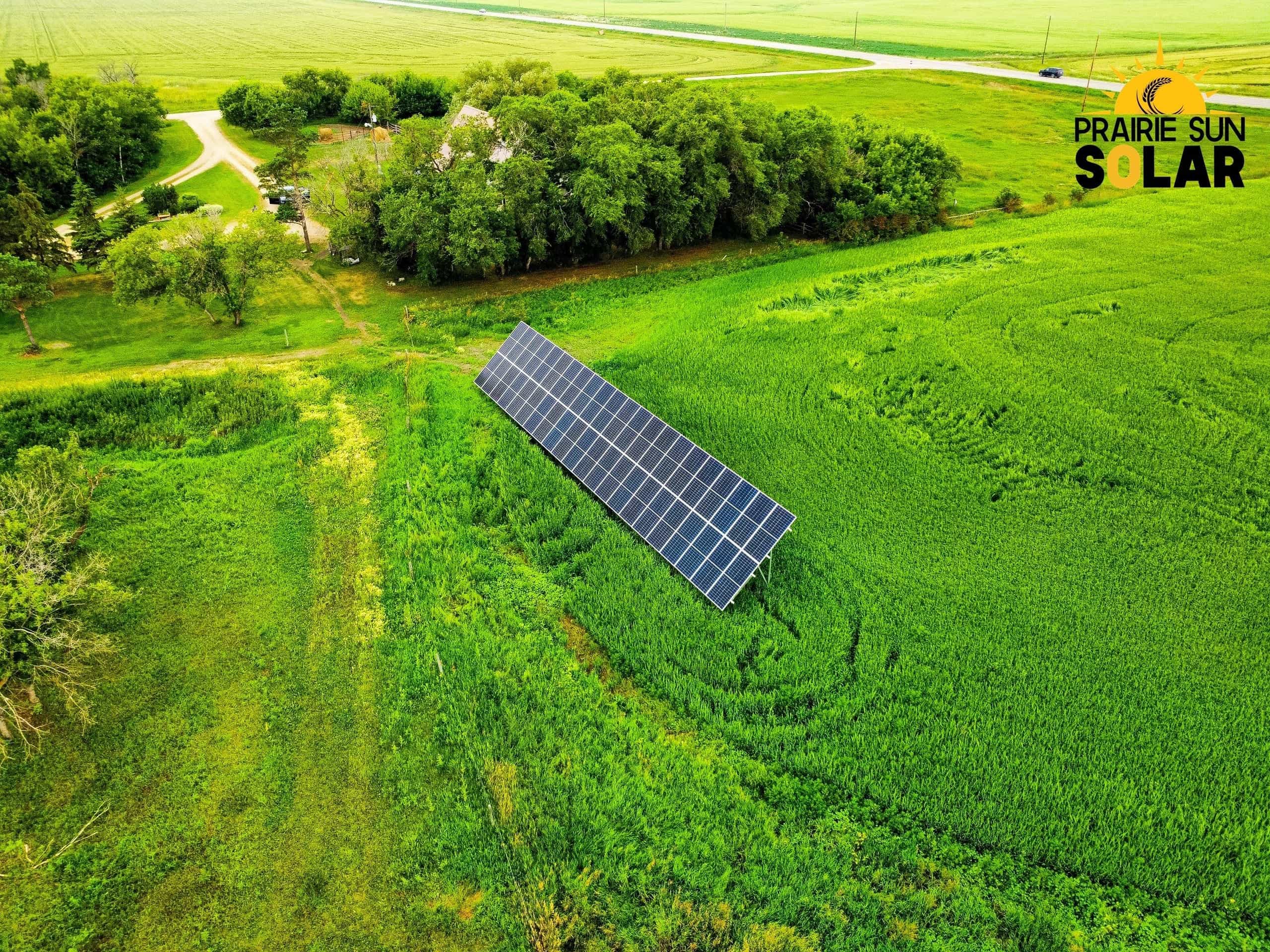 Aerial view of a single long row of ground-mount solar panels in a green hayfield, with the Audette farmyard and shelterbelt behind