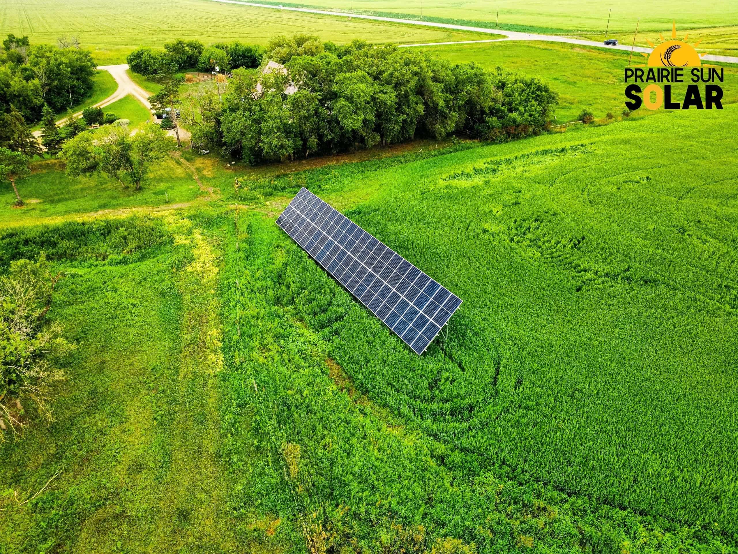 Aerial view of a single long row of ground-mount solar panels in a green hayfield, with the Audette farmyard and shelterbelt behind
